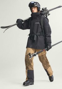 Crna vodootporna jakna s logom, smešane hlače s uzorkom, skijaške čizme, kaciga i skije, sa detaljima kao što su kaiševi i skijaški štapovi. Glatka tekstura.