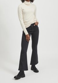 Maglione a collo alto beige chiaro con maniche lunghe, abbinato a jeans neri a vita alta a campana e stivali neri alla caviglia. Texture morbide.