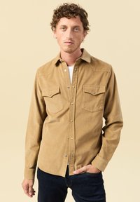 Giovane con capelli ricci che indossa una camicia di velluto a coste color beige sopra una maglietta bianca e jeans scuri, in piedi davanti a uno sfondo beige.