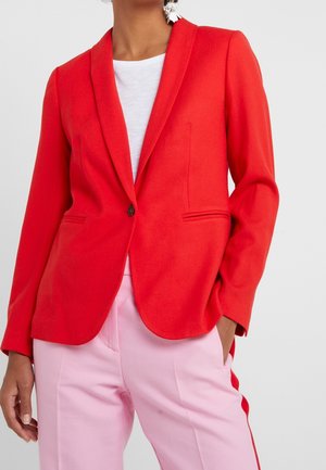 Blazer rouge en tissu lisse avec un col à revers, fermeture à un bouton, et deux poches latérales, associé à un pantalon rose clair.