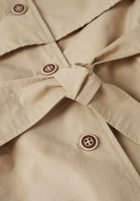 Beżowy płaszcz trench z wiązanym w pasie, z brązowymi guzikami i ostrym kołnierzem. Gładka tekstura z czystym, dopasowanym krojem.