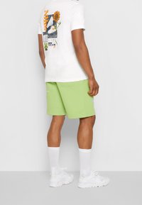 Camiseta de algodón blanca con estampado gráfico multicolor en la parte posterior, combinada con pantalones cortos de algodón verdes y zapatillas blancas de diseño voluminoso.