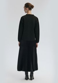 Musta pitkähihaine paita, joka on yhdistetty pleated mustaan hameeseen, jossa on sileä pinta ja kerroksellinen ilme, täydentäen asukokonaisuutta korkeakorkoisilla saappailla.