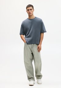 Šedé oversized tričko s krátkými rukávy, spárované s bledě zelenými volnými džíny a bílými teniskami, na čistém bílém pozadí.