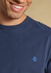 Lächelnde Person trägt ein dunkelblaues T-Shirt mit Rundhalsausschnitt und einem kleinen hellblauen bestickten Logo auf der Brust.