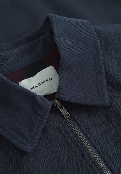Navyblauwe zip-up jas met een gestructureerde ribbelkraag en een gladde stof, met een zichtbare rits en een label waarop "WOOD WOOD" staat.