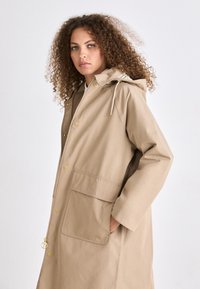 Beige waterdichte jas met capuchon, voorzien van gouden hardware, twee grote voorzakken en een ritsluiting. Glad textuur, middellange ontwerp.
