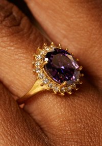 Goldring mit einem ovalen lila Edelstein, umgeben von kleineren klaren Steinen. Der Ringdraht hat ein strukturiertes Design mit gefassten Steinchen.