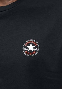 Μαύρο φούτερ με στρογγυλό, λευκό λογότυπο που περιλαμβάνει ένα αστέρι, το κείμενο "CONVERSE" και "All Star" με κόκκινα γράμματα, και το "Chuck Taylor" με μαύρα γράμματα. Μαλακή υφή.