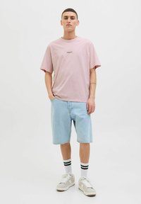 Lyserød bomulds T-shirt med korte ærmer, parret med lyseblå denimshorts. Modellen bærer hvide sneakers med sorte stribede sokker.