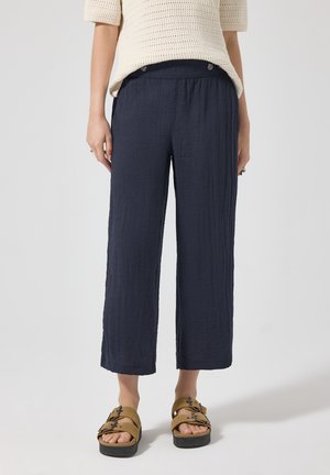 Kvinde iført cropped navy viddebenede bukser, beige kortærmet åbenvævet top og brune sandaler med spænder, stående på en lys baggrund.