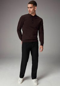 Camisa polo de manga larga color marrón oscuro con tres botones, combinada con pantalones negros y zapatillas blancas. Textura suave, diseño ajustado.