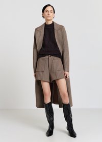 Cappotto marrone in misto lana con ampi risvolti, maglione scuro lavorato a maglia, pantaloncini marroni su misura abbinati e stivali al ginocchio in pelle verniciata nera.
