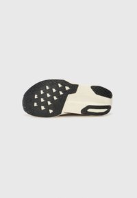 La semelle de la chaussure présente une surface en caoutchouc noir texturé avec un motif de crampons triangulaire et une semelle intermédiaire de couleur crème. Décorée d'une découpe visible.