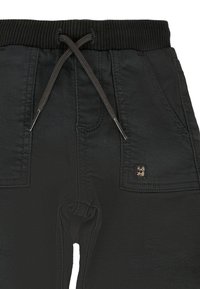 Pantalones de chándal negros con cintura ajustable mediante cordón, banda elástica, dos bolsillos laterales y un pequeño parche con logo cerca del bolsillo. Textura de tela suave.