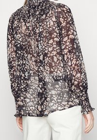 Blouse noire en transparence avec un col froncé, motif floral en beige et bleu, manches longues avec des poignets élastiques, et tissu fluide.