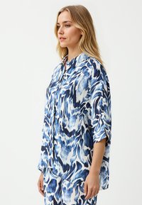 Bluse in Blau und Weiß mit Muster, lässiger Schnitt, Kragen und kurzen Ärmeln. Der Stoff wirkt leicht und leicht strukturiert.