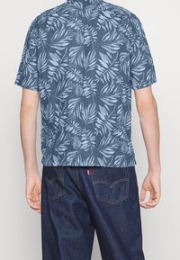 Camicia a maniche corte in tessuto blu scuro con un motivo di foglie tropicali blu chiaro, dotata di colletto con bottoni e orlo dritto.