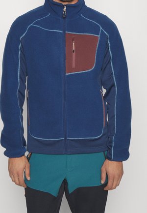 Mężczyzna w niebieskiej polarowej kurtce z jasnoniebieskimi przeszyciami i brązową kieszenią na piersi na zamek, w zestawie z zielonkawymi i czarnymi spodniami.