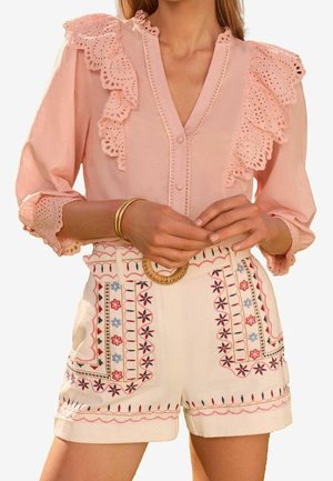 Blouse rose clair avec bordure en dentelle ajourée à volants, boutonnée devant, manches trois-quarts. Associée à un short crème brodé avec des motifs floraux.