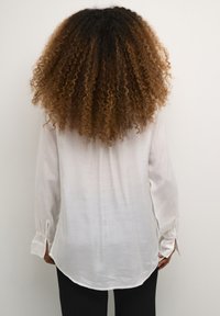 Blusa bianca a maniche lunghe realizzata in tessuto leggero con orlo curvo e dettagli ai polsini, indossata con pantaloni neri.
