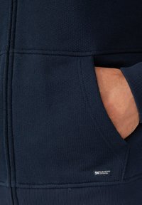Felpa con zip di un blu navy scuro realizzata in tessuto testurizzato, con polsini e fondo a costine, dotata di una tasca frontale e di un piccolo logo nella parte inferiore.
