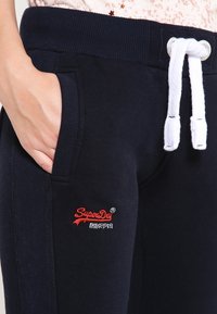 Marinblå joggers med ribbat midjeband, vit dragsko, sidofickor och röd broderad logotyp på vänster lår. Mjuk materialkänsla.