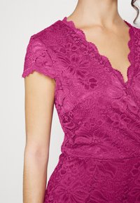 Vestido de renda fúcsia com mangas curtas e recortadas, decote em V profundo, padrões florais e acabamento texturizado. Close-up da área superior do ombro.