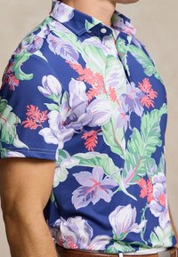 Camicia a maniche corte con uno sfondo blu e grandi motivi floreali in rosa, verde e viola; include un colletto con bottoni.