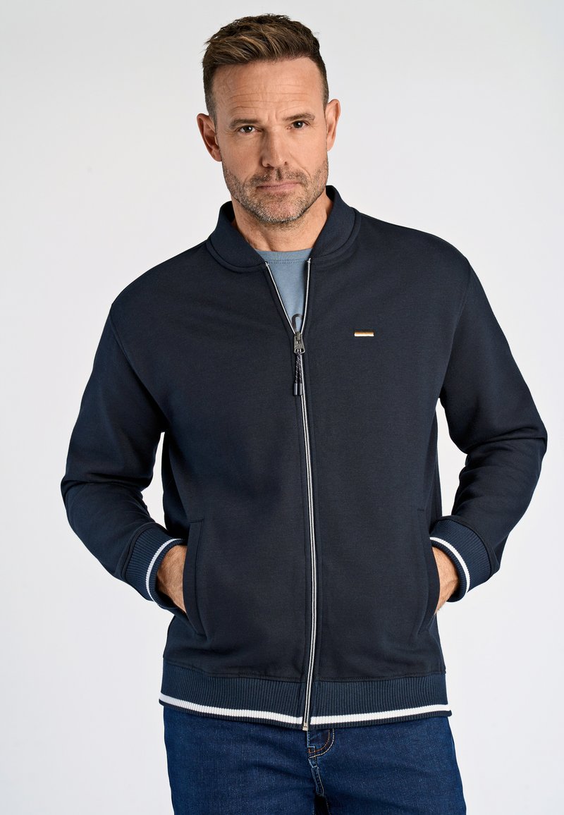 Giacca a zip navy realizzata in materiale morbido; presenta polsini e orlo a costine con strisce azzurre e bianche; include un piccolo logo decorativo.