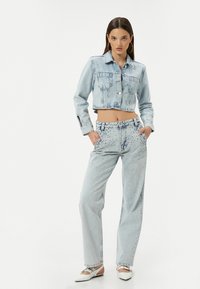 Chaqueta corta de mezclilla azul claro con cierre de botones y dos bolsillos en el pecho, combinada con pantalones vaqueros de pierna ancha azul claro que presentan detalles de tachuelas.
