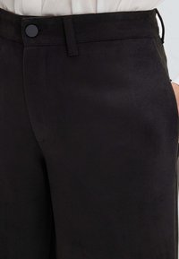 Schwarze Hose aus einem glatten Stoff. Verfügt über eine flache Vorderseite, einen Knopfverschluss und seitliche Taschen. Saubere Linien und minimale Details.