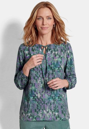 Femme aux cheveux blonds mi-longs portant un chemisier à manches longues violet et vert à motifs avec un lien au niveau de l'encolure.