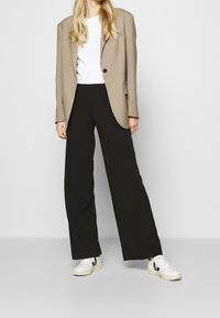 Beige blazer med en enkelt sort knap, parret med sorte vide bukser og hvide sneakers med sorte detaljer, stående på en neutral baggrund.