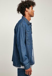 Denim jakas vidējā zilā krāsā ar brīvu piegriezumu, ar garām piedurknēm, pogu aizdari un krūšu kabatu. Gluda tekstūra.