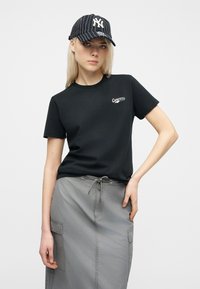 Νεαρό άτομο φορώντας μαύρο μπλουζάκι Converse, γκρι φούστα cargo και μαύρο καπέλο New York Yankees με ρίγες, σε λευκό φόντο.