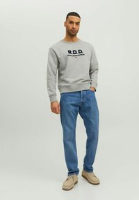 Grauer Hoodie mit "R.D.D." und "ROYAL DENIM DIVISION" in Schwarz gedruckt. Getragen mit blauen Jeans und beigen Schuhen. Einfaches Design und entspannter Schnitt.