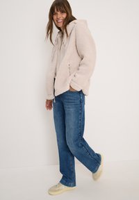 Femme souriante, portant une veste en polaire crème, un sweat à capuche léger, un jean bleu et des baskets beiges, se tenant devant un fond clair uni.