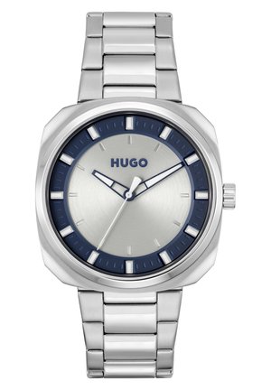 Montre en acier inoxydable argenté avec une lunette bleu foncé, un cadran argenté, des index d'heure blancs et un bracelet à maillons métalliques. Présente un design simple et moderne.