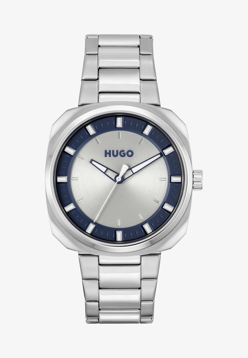 Reloj de acero inoxidable plateado con un bisel azul oscuro, esfera plateada, marcadores de hora blancos y una pulsera de eslabones de metal. Presenta un diseño simple y moderno.