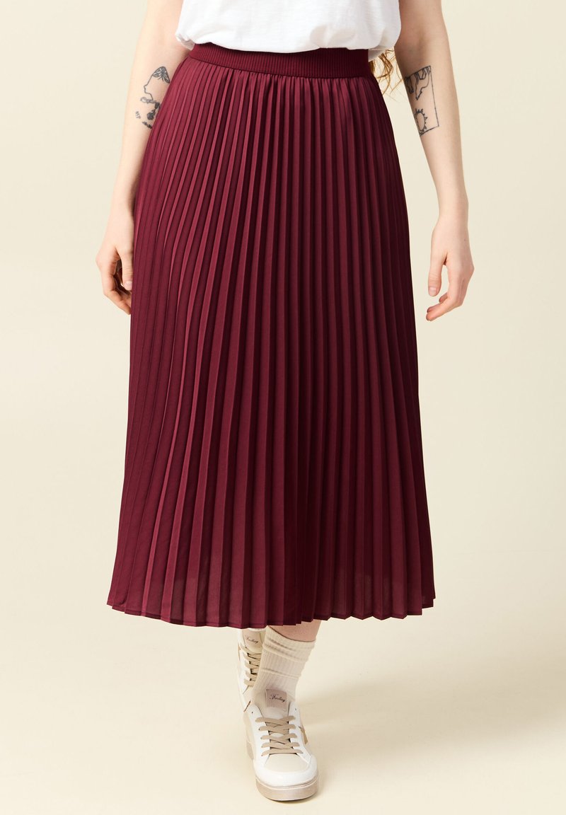 Burgunderröd plisserad midikjol med mjuk elastisk midja. Buren med vita sneakers och ljusa strumpor, vilket visar en flödande design.