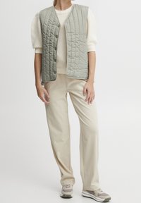 Gilet matelassé gris clair avec une coupe décontractée, doté de boutons-pression et associé à un pull en tricot côtelé en dessous, le tout complété par un pantalon beige et des baskets.