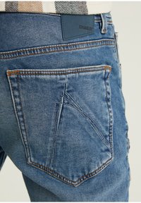 Džínové kalhoty v světle modré barvě s vybledlým efektem. Mají kapsu ve tvaru trojúhelníku a kontrastní prošívání. Kožená značka značky na pásu.