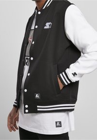 Veste universitaire noire et blanche avec boutons-pression, poignets côtelés, poches latérales et écussons logo. Présente des manches contrastantes et des rayures.