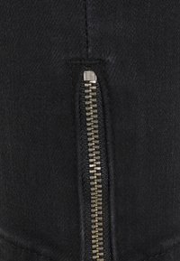 Tejido de mezclilla negra con un detalle de cremallera de metal en el costado. La textura parece suave con un leve brillo en el hardware de la cremallera.