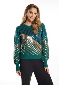 Groene trui met glinsterende pailletten in diagonale strepen van brons en blauw. Heeft een ronde halslijn en lange, opgeblazen mouwen.