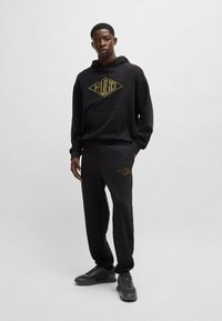 Felpa nera con cappuccio e pantaloni jogger con un logo dorato sul petto e sulla gamba. Miscelato in cotone, vestibilità rilassata, cappuccio regolabile e tasca frontale. Sneakers nere.