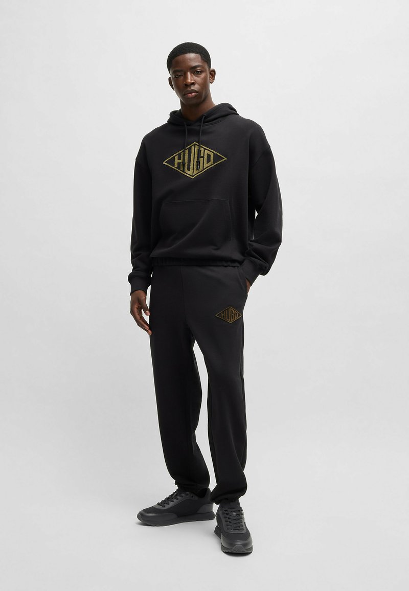 Felpa nera con cappuccio e pantaloni jogger con un logo dorato sul petto e sulla gamba. Miscelato in cotone, vestibilità rilassata, cappuccio regolabile e tasca frontale. Sneakers nere.
