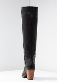Botte haute en cuir noir avec une finition texturée, une forme effilée et un talon en bois. Des coutures décoratives courent le long du côté.