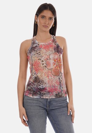 Mujer con cabello largo y oscuro, lleva pendientes de aro dorados, una blusa sin mangas con estampado floral colorido y jeans azules, de pie contra un fondo sencillo.